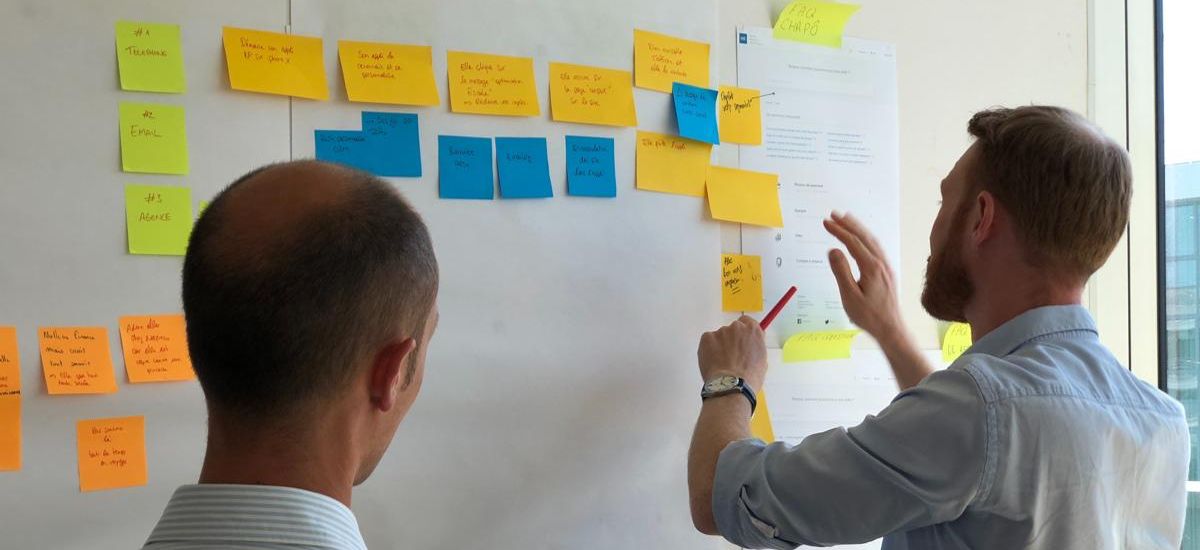 Mann steht vor weißer Wand mit bunten Postits und wird von weiterem Mann beobachtet.