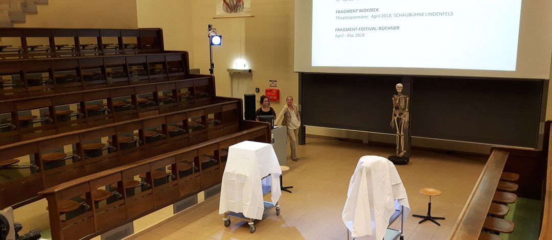 Blick in einen Anatomiehörsaal mit halbrunder Bestuhlung; in der Mitte stehen ein Skelett und zwei mit weißen Tüchern verhüllte Transportwägen