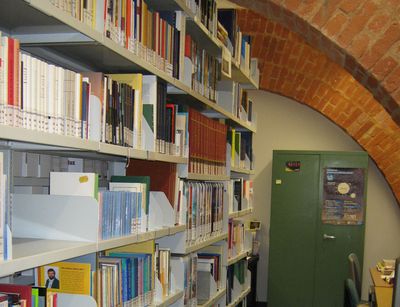 Links im Bild nebeneinander aufgereihte Bücherregale. Gegenüber Schreibtische mit Bürostühlen.